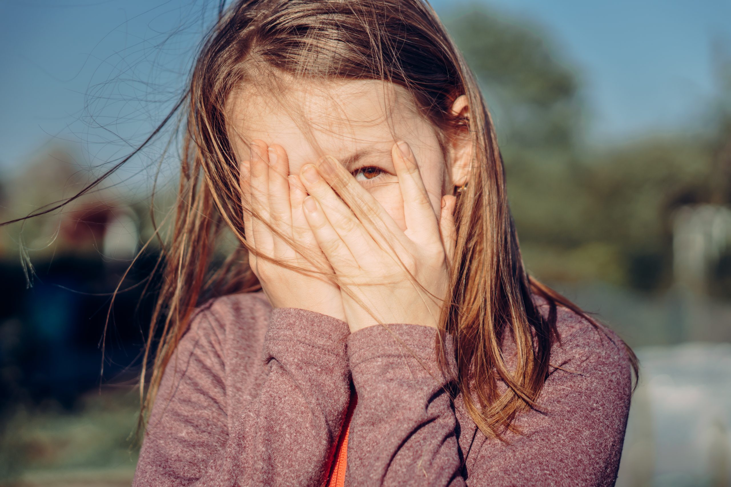 Meisje met lang haar verbergt haar gezicht met beide handen terwijl ze door haar vingers kijkt, buiten in het zonlicht.