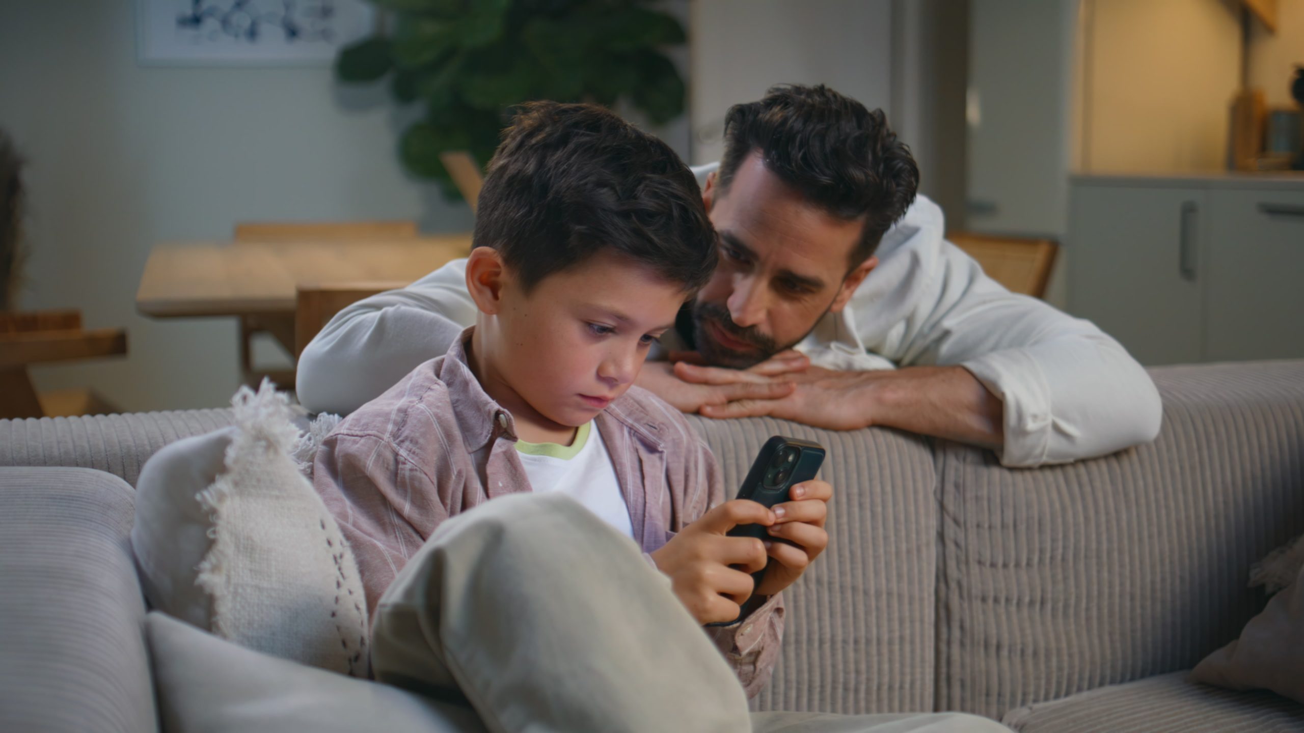 Vader kijkt bezorgd naar zijn zoon die geconcentreerd op zijn smartphone zit, zittend op de bank in de woonkamer.