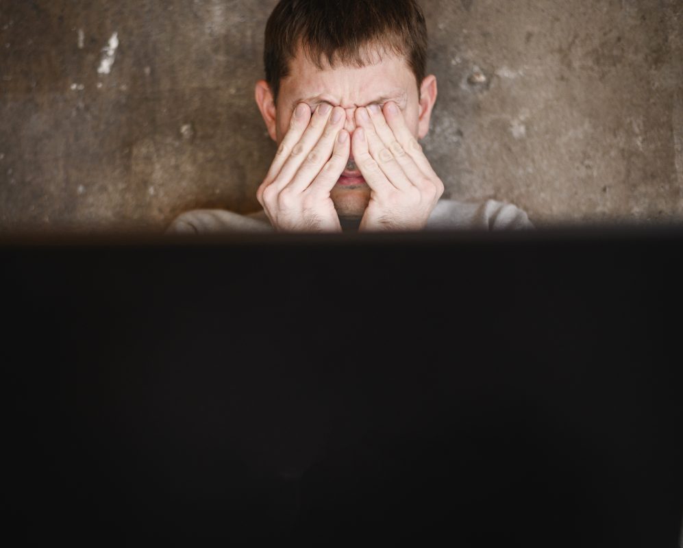 Jongere met gesloten ogen en handen op het gezicht voor een computerscherm, zichtbaar worstelend met emotionele stress.
