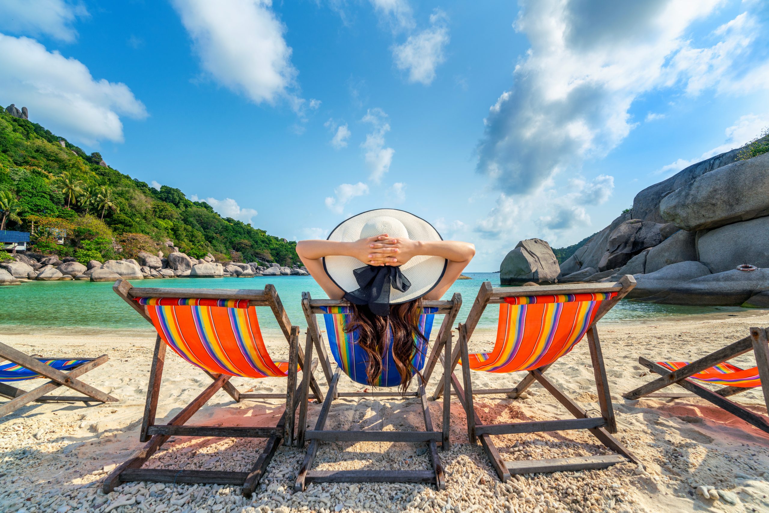 Vrouw met zonnenhoed ontspant op kleurrijke strandstoel aan tropisch strand met helderblauw water en palmbomen.