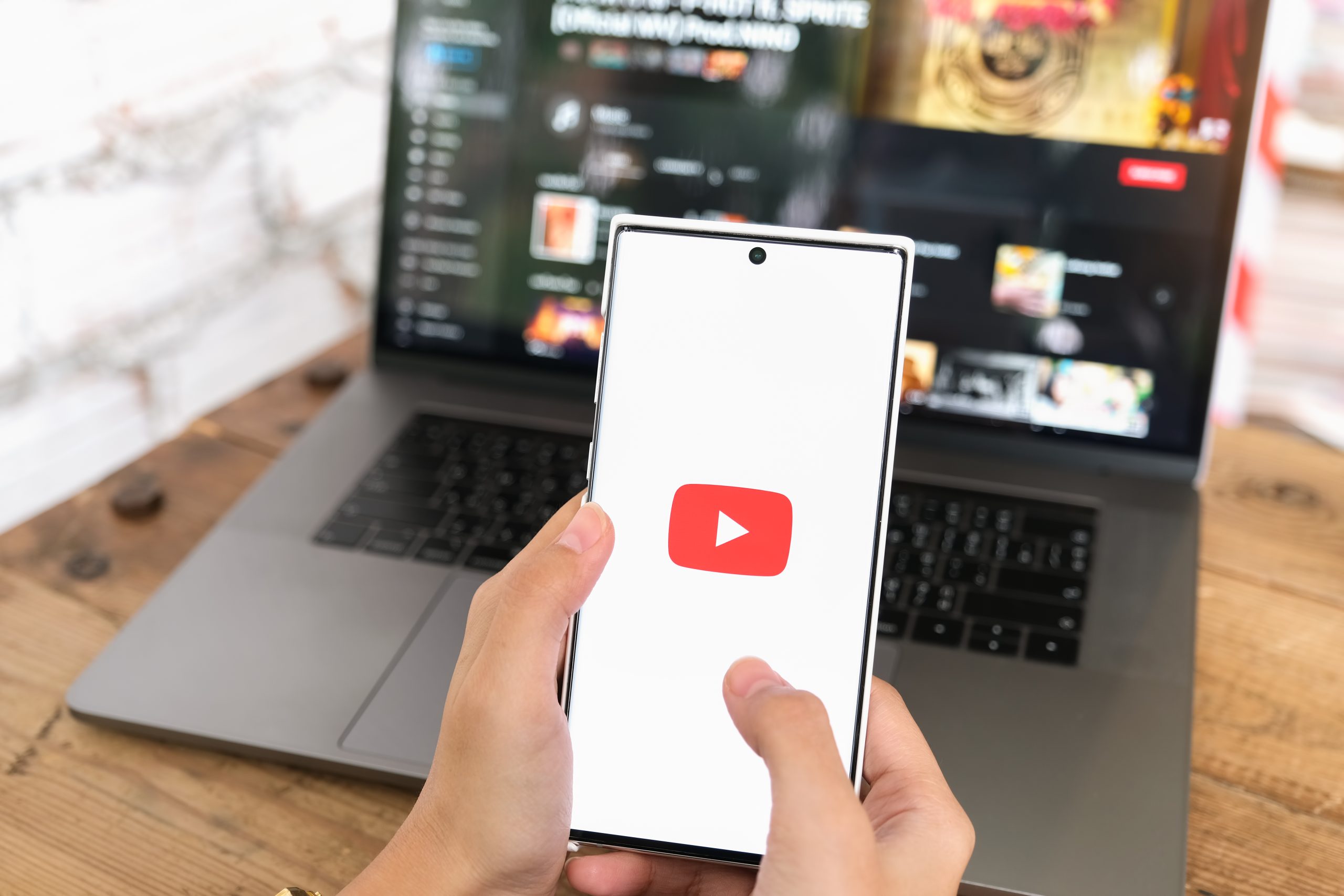 Persoon houdt smartphone vast met YouTube-logo op het scherm, terwijl op de achtergrond een laptop met geopende YouTube-pagina staat.