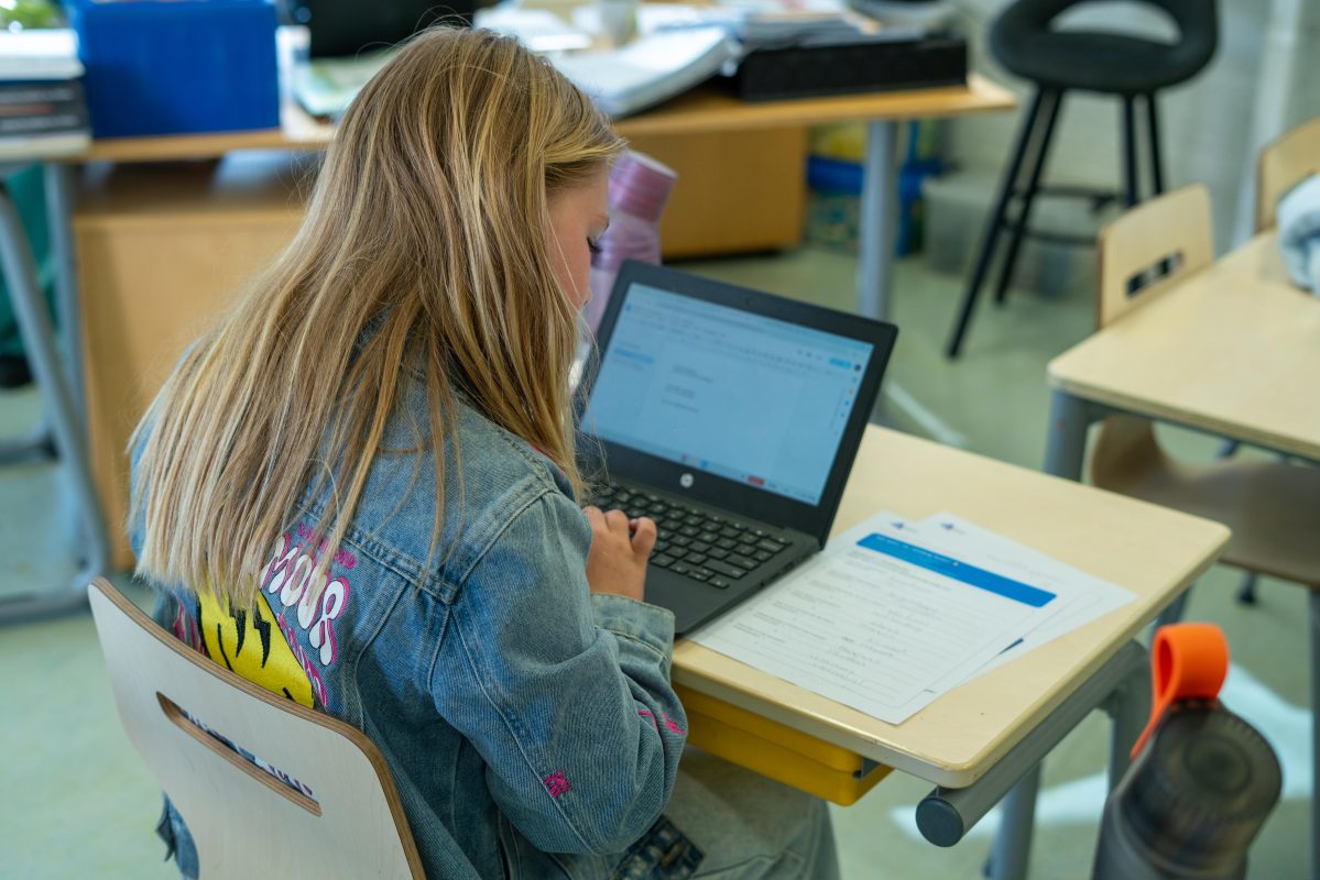 Meisje in klaslokaal typt op een laptop terwijl een werkblad op haar bureau ligt