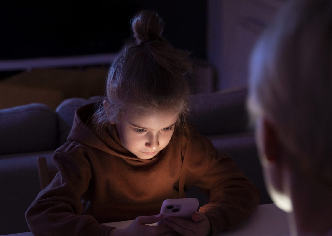 Kind zit ’s avonds verdiept in smartphone, terwijl ouder toekijkt aan tafel.