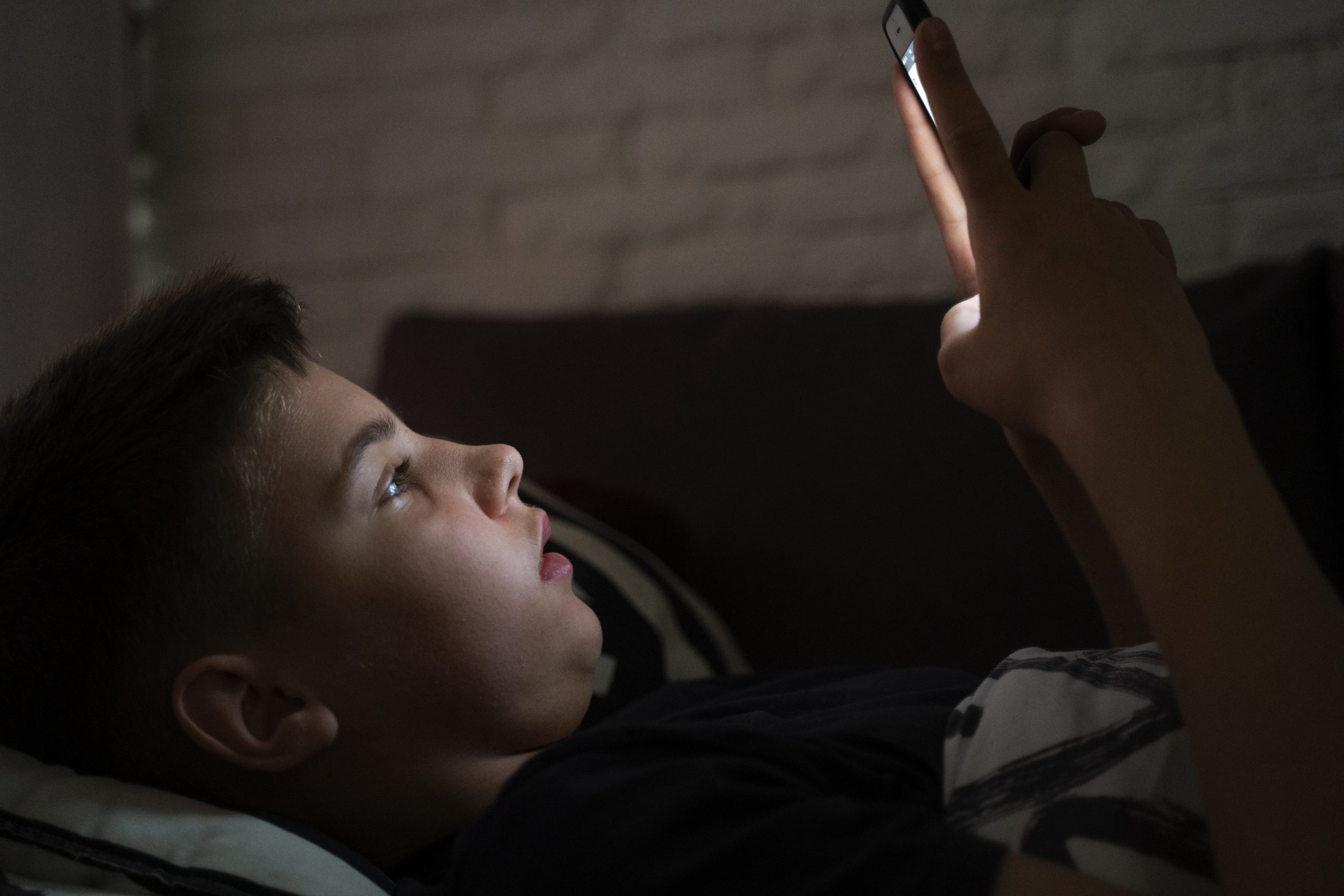 Jonge jongen ligt in bed en kijkt in het donker op zijn smartphone, met blauw licht op zijn gezicht