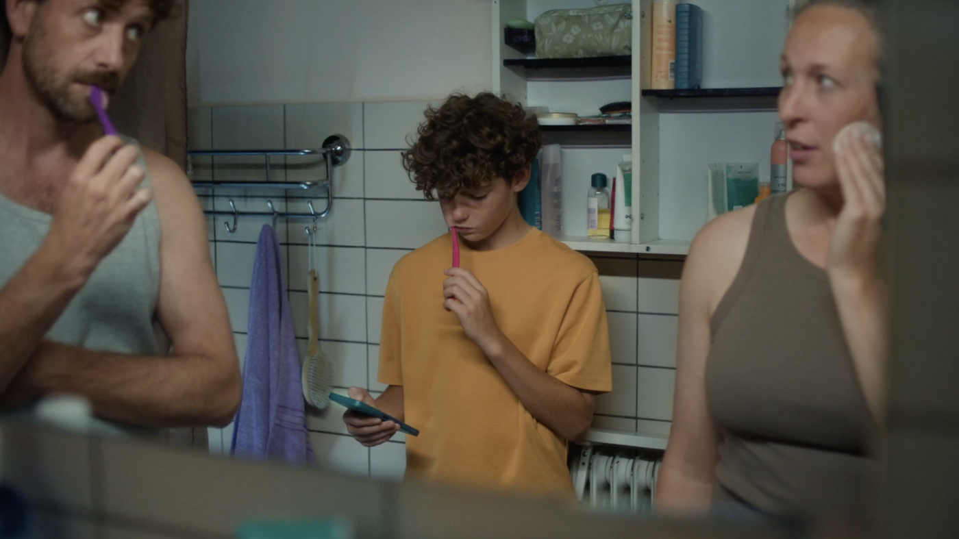 Jongen kijkt op smartphone tijdens tandenpoetsen, terwijl ouders erbij staan in de badkamer.