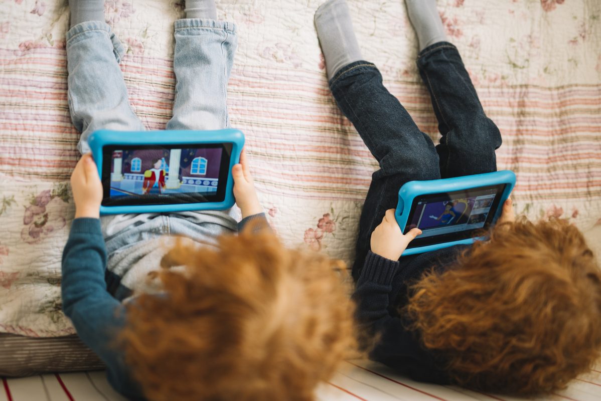 Twee jonge kinderen liggen op bed met een tablet in hun handen en spelen een online spel.