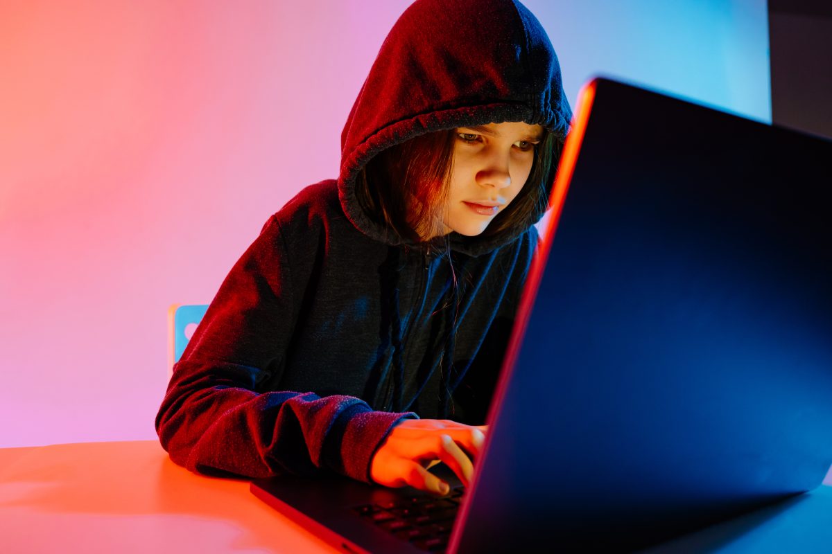 Jongere met hoodie typt geconcentreerd op laptop in kleurrijke belichting.