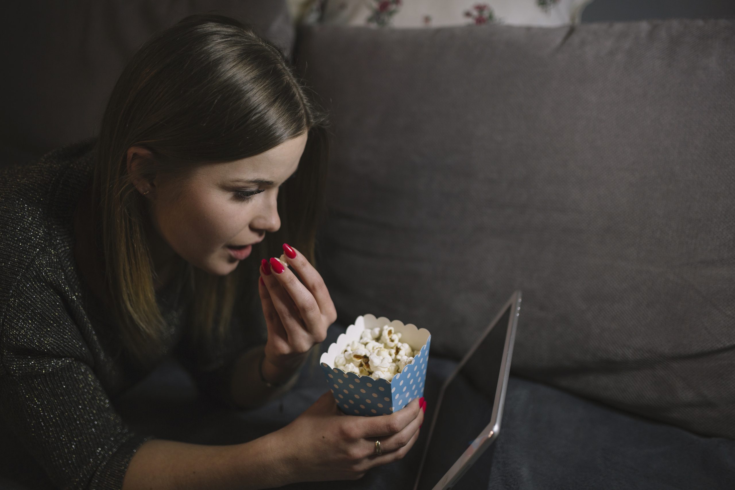 Jonge vrouw die ontspannen op de bank popcorn eet terwijl ze een video kijkt op een tablet.