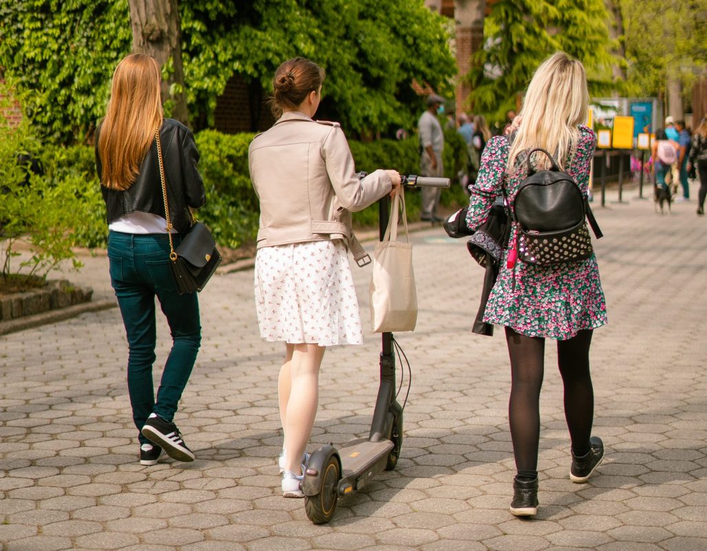 Drie jonge vrouwen lopen over een zonnig parkpad, waarvan één een elektrische step duwt.