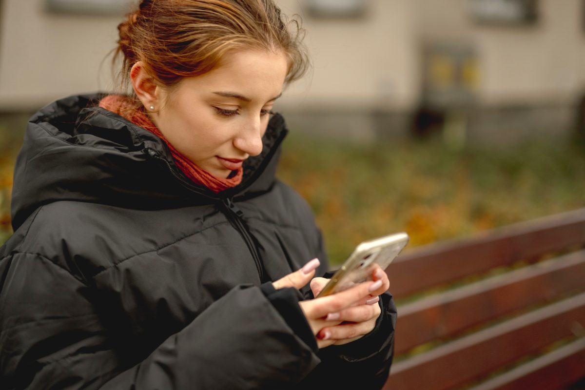 Jongere kijkt geconcentreerd op smartphone terwijl zij buiten op een bankje zit