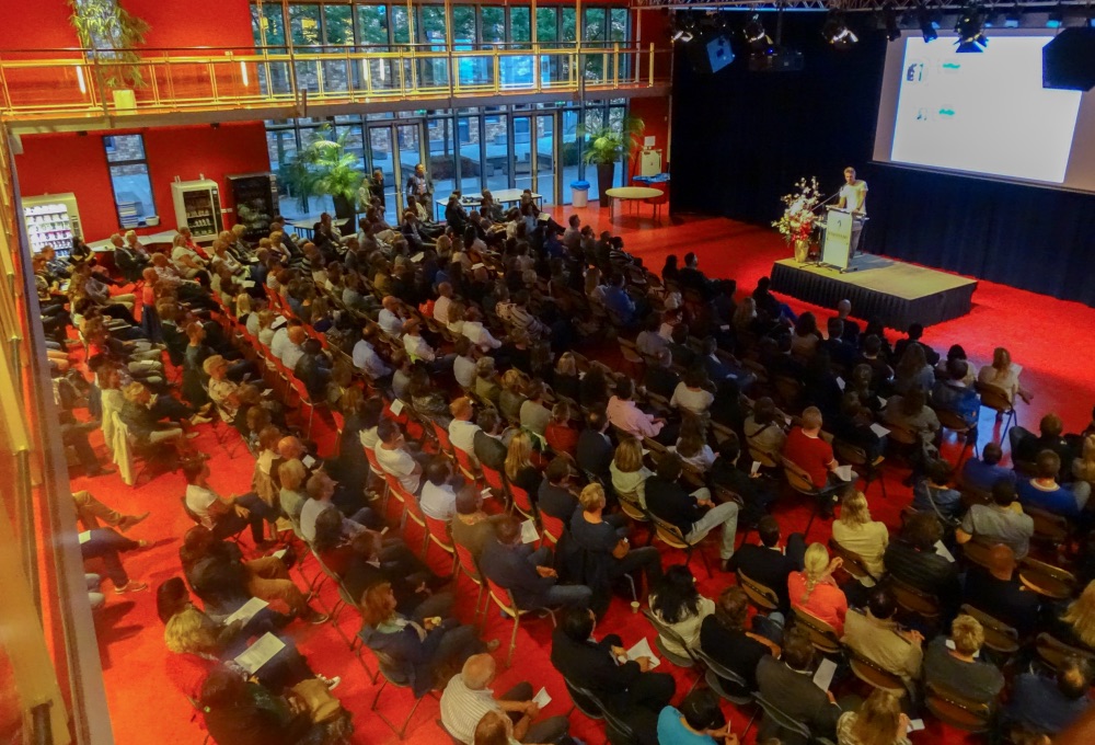 Ouders zitten in een zaal tijdens een ouderavond over digitale geletterdheid op een school terwijl een spreker een presentatie geeft op een podium.