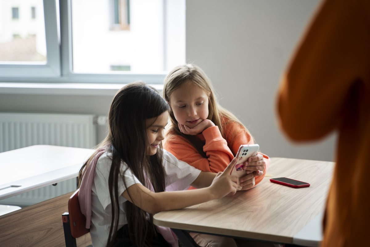 Twee jonge leerlingen kijken samen naar een smartphone in de klas terwijl een docent op de achtergrond staat