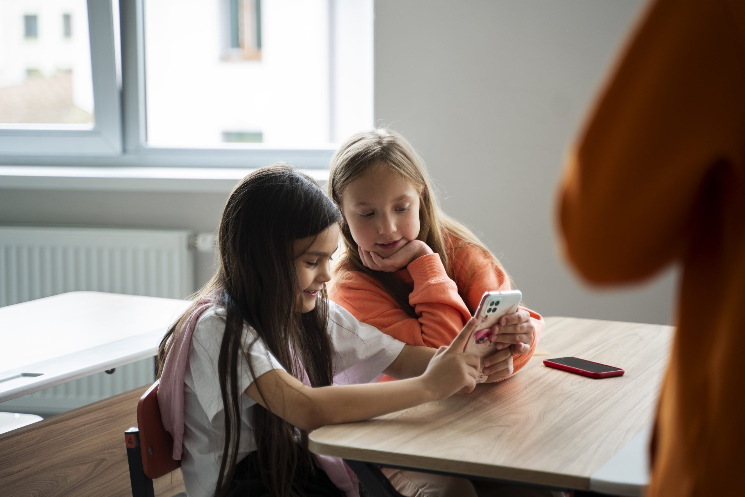 Twee jonge leerlingen kijken samen naar een smartphone in de klas terwijl een docent op de achtergrond staat
