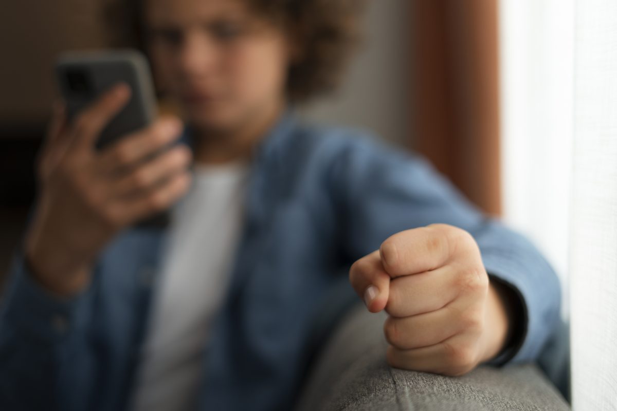 Jongere kijkt boos en gefrustreerd naar zijn telefoon terwijl hij een gebalde vuist maakt, passend bij afwijzing en negatieve online ervaringen