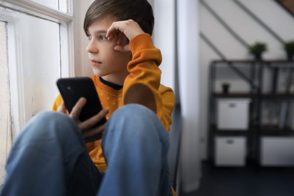 Jongen kijkt bezorgd naar zijn telefoon terwijl hij bij het raam zit.