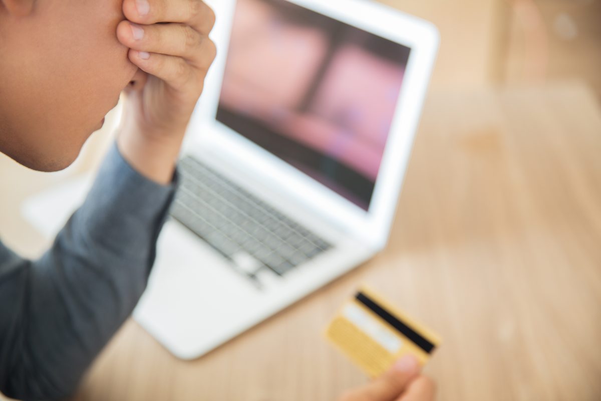 Jongen kijkt gestrest naar een creditcard terwijl op de laptop een onscherpe online omgeving zichtbaar is, passend bij online fraude en digitale oplichting.