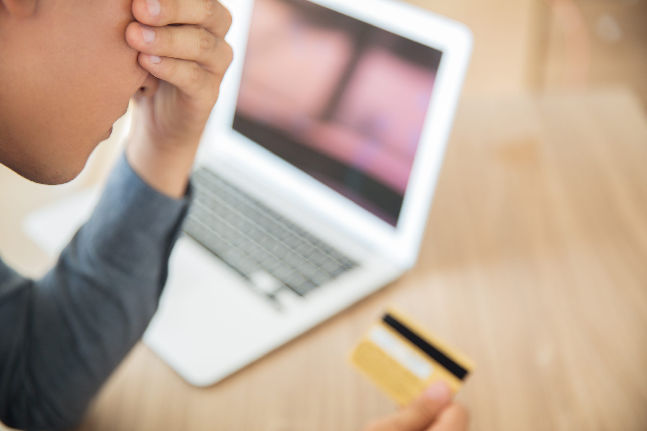 Jongen kijkt gestrest naar een creditcard terwijl op de laptop een onscherpe online omgeving zichtbaar is, passend bij online fraude en digitale oplichting.