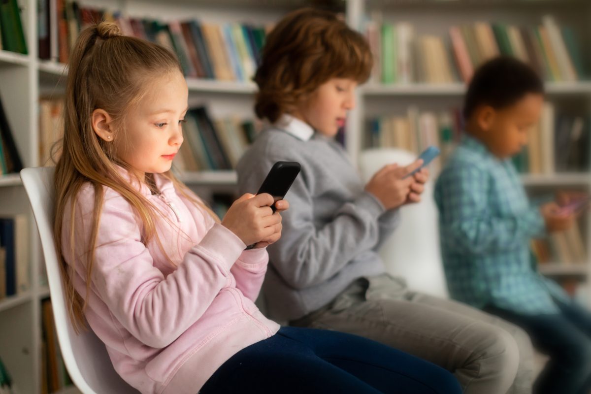 Drie basisschoolkinderen zitten in een klaslokaal en kijken op hun smartphone tijdens een pauzemoment.