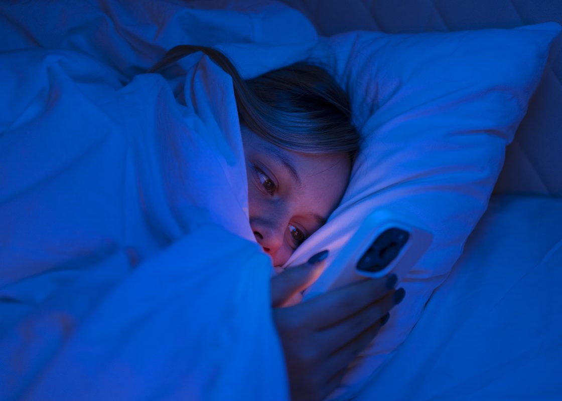 Jongere ligt ’s nachts in bed op haar telefoon te scrollen, passend bij doomscrolling en overmatig socialmediagebruik.