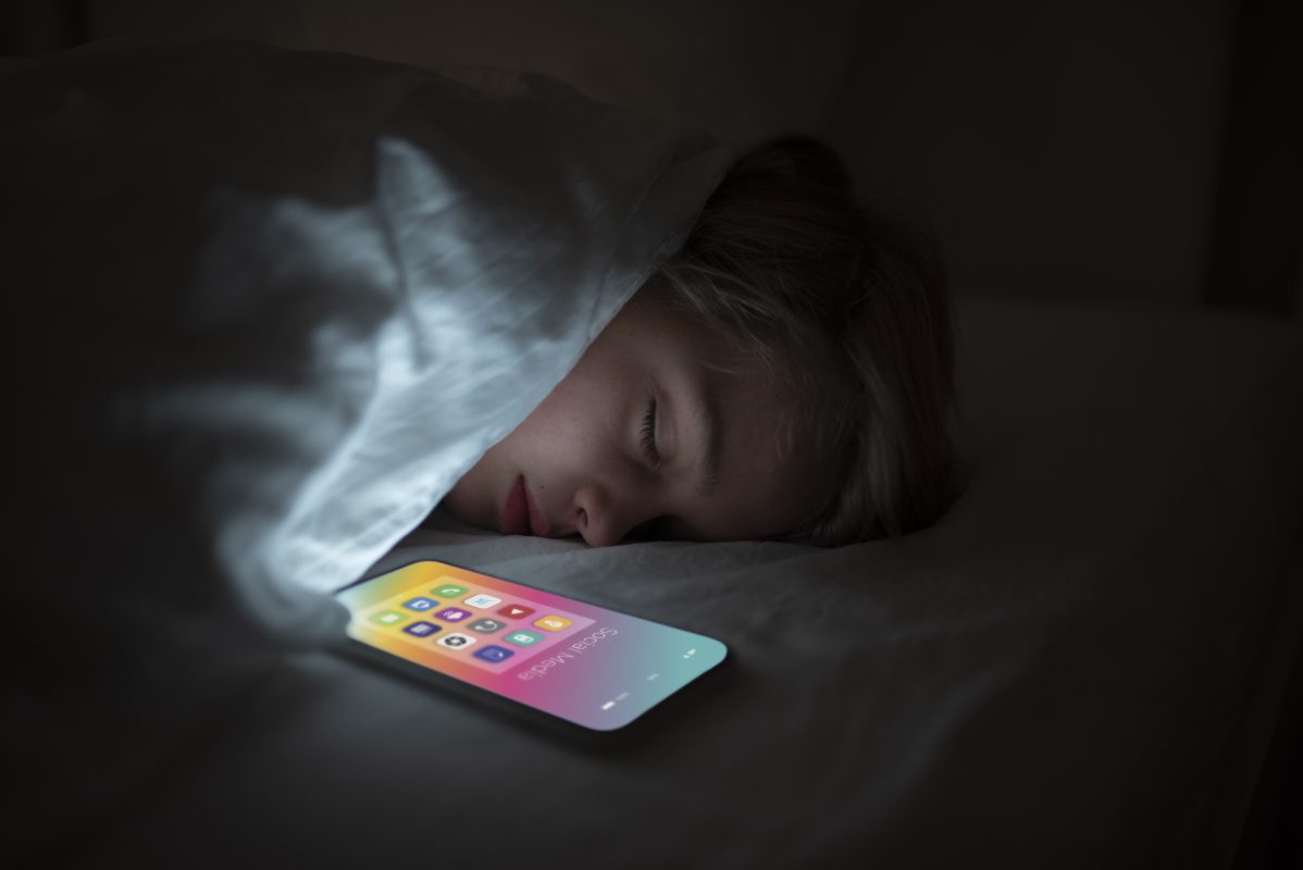 Jong meisje ligt 's avonds in bed terwijl het licht van een smartphone haar gezicht verlicht, als illustratie van intensief socialmediagebruik en de invloed van schermen op slaap en welzijn.