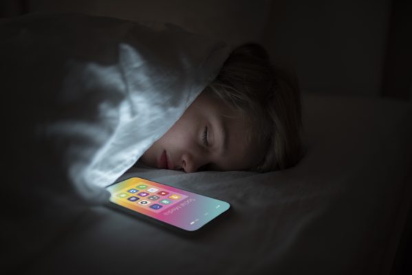 Jong meisje ligt 's avonds in bed terwijl het licht van een smartphone haar gezicht verlicht, als illustratie van intensief socialmediagebruik en de invloed van schermen op slaap en welzijn.