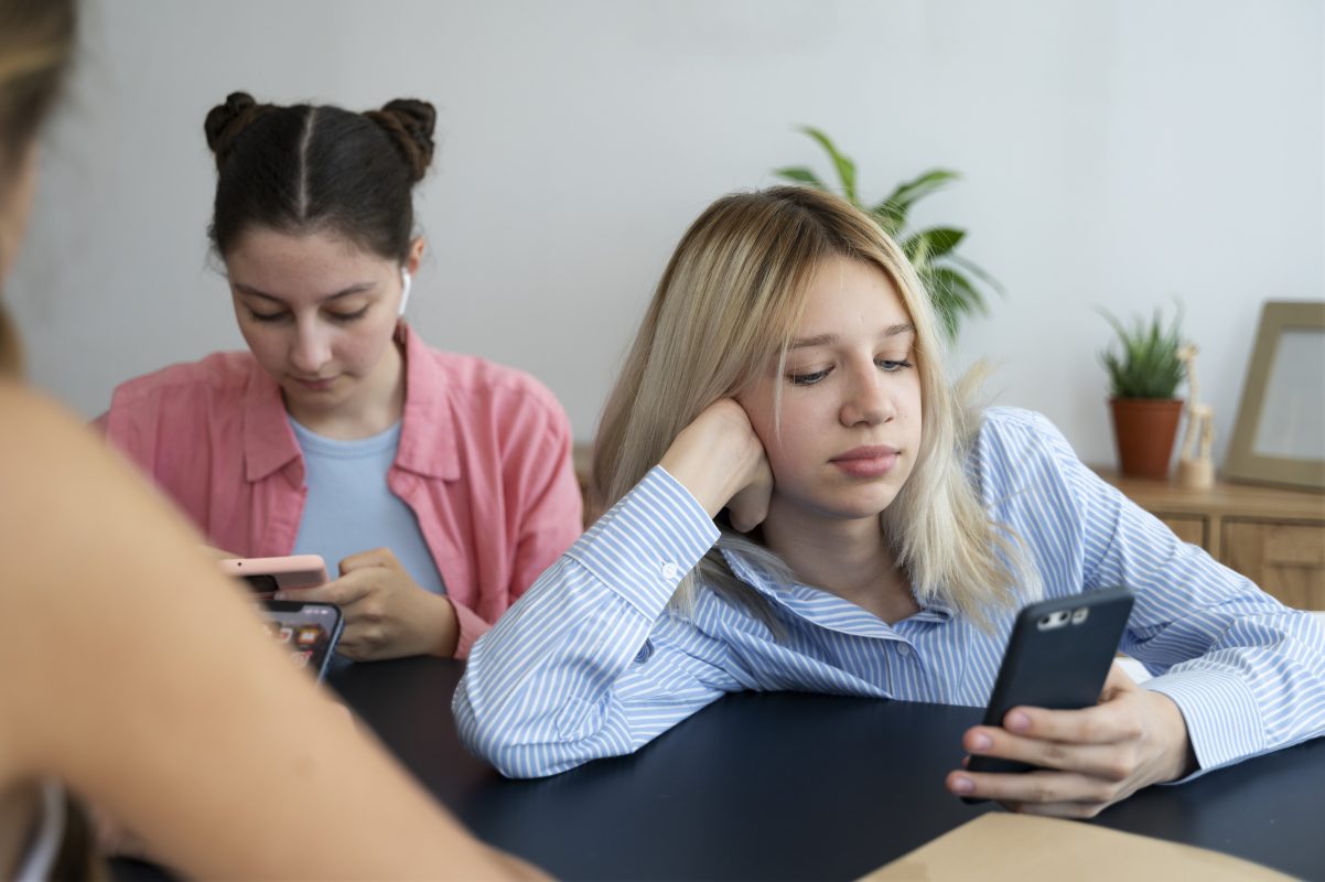 Twee tieners kijken op hun smartphone terwijl ze samen aan tafel zitten, passend bij de impact van social media op mentale gezondheid bij jongeren.