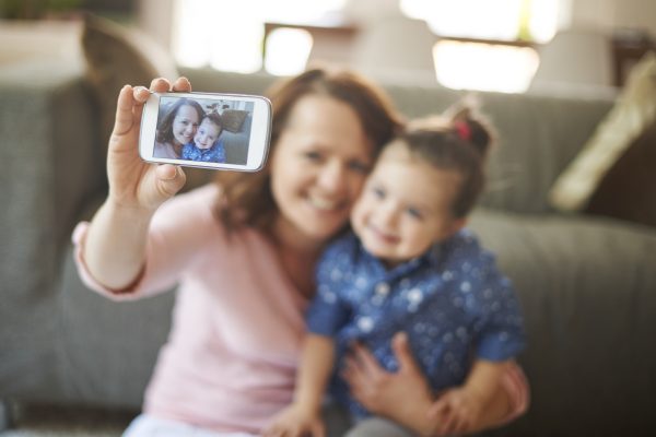 Moeder maakt samen met haar jonge dochter een selfie met een smartphone, passend bij de discussie over kinderen in vlogs, sociale media en online zichtbaarheid.