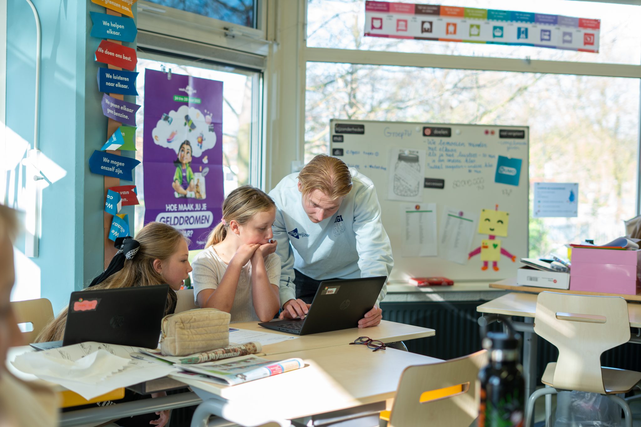Een Digiwijzer-digicoach helpt twee leerlingen tijdens een les digitale geletterdheid terwijl zij samen op een laptop werken.