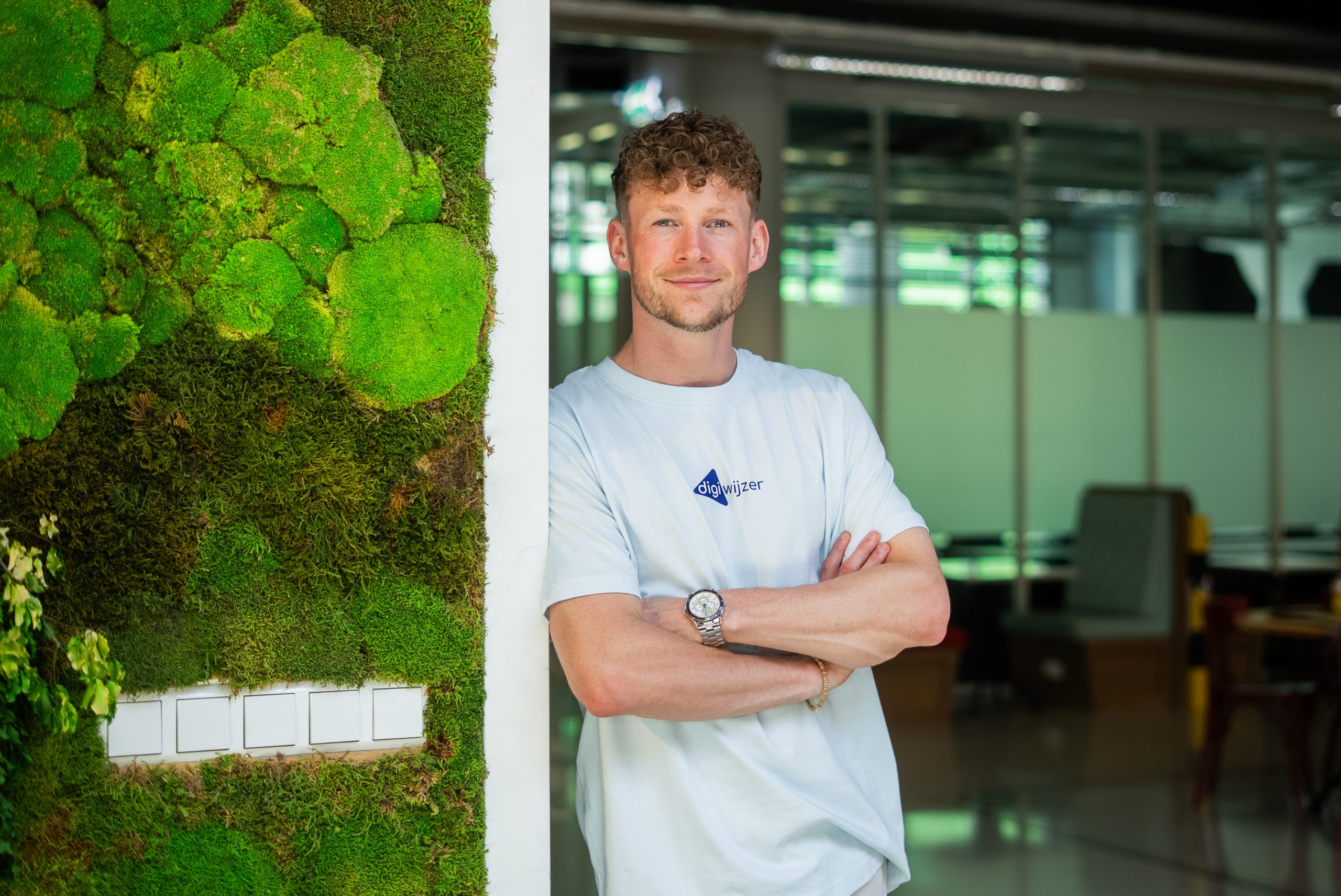 Portret van een Digiwijzer-medewerker die met gekruiste armen naast een groene moswand staat op het kantoor van Digiwijzer.