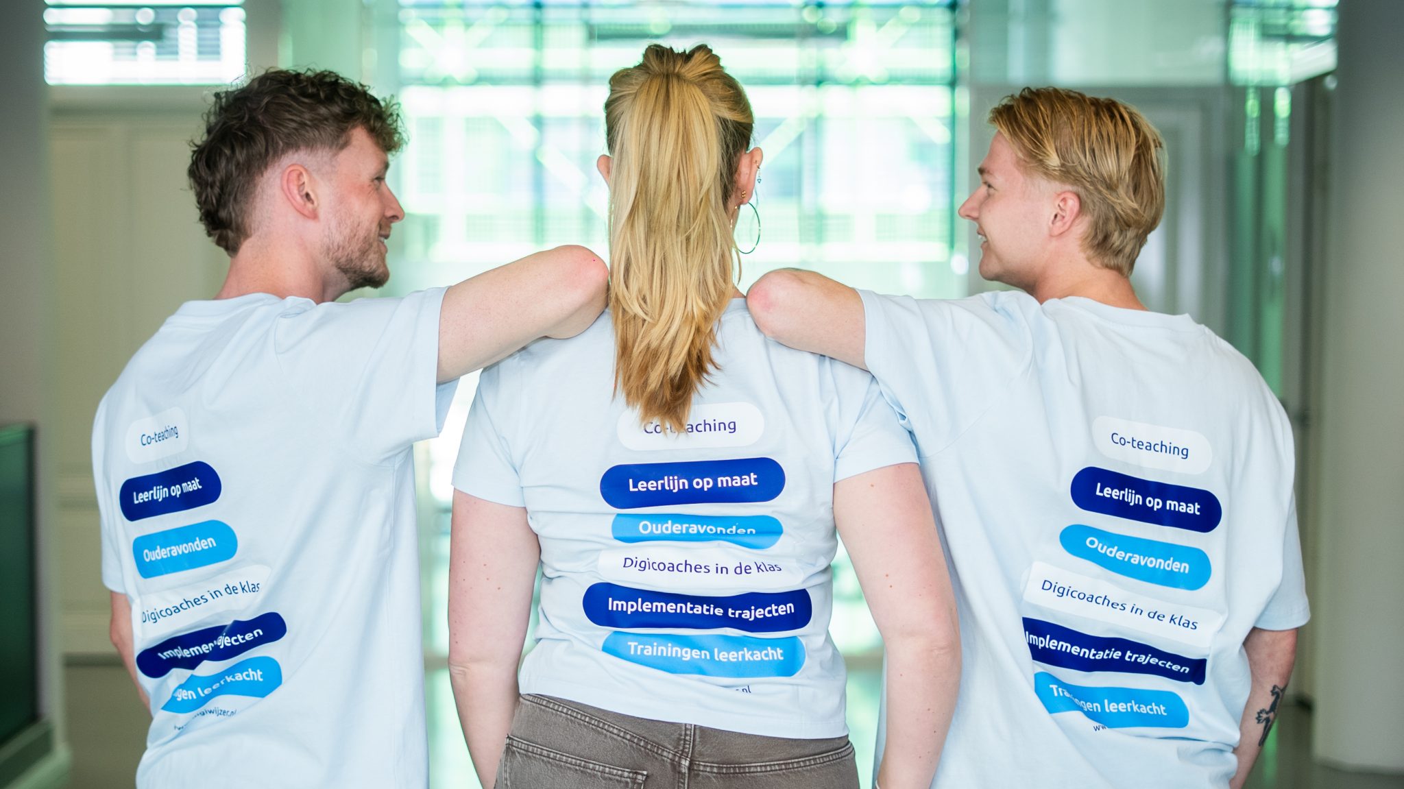 Drie medewerkers van Digiwijzer staan met hun armen over elkaars schouders en tonen de verschillende oplossingen voor digitale geletterdheid op de achterkant van hun shirts.