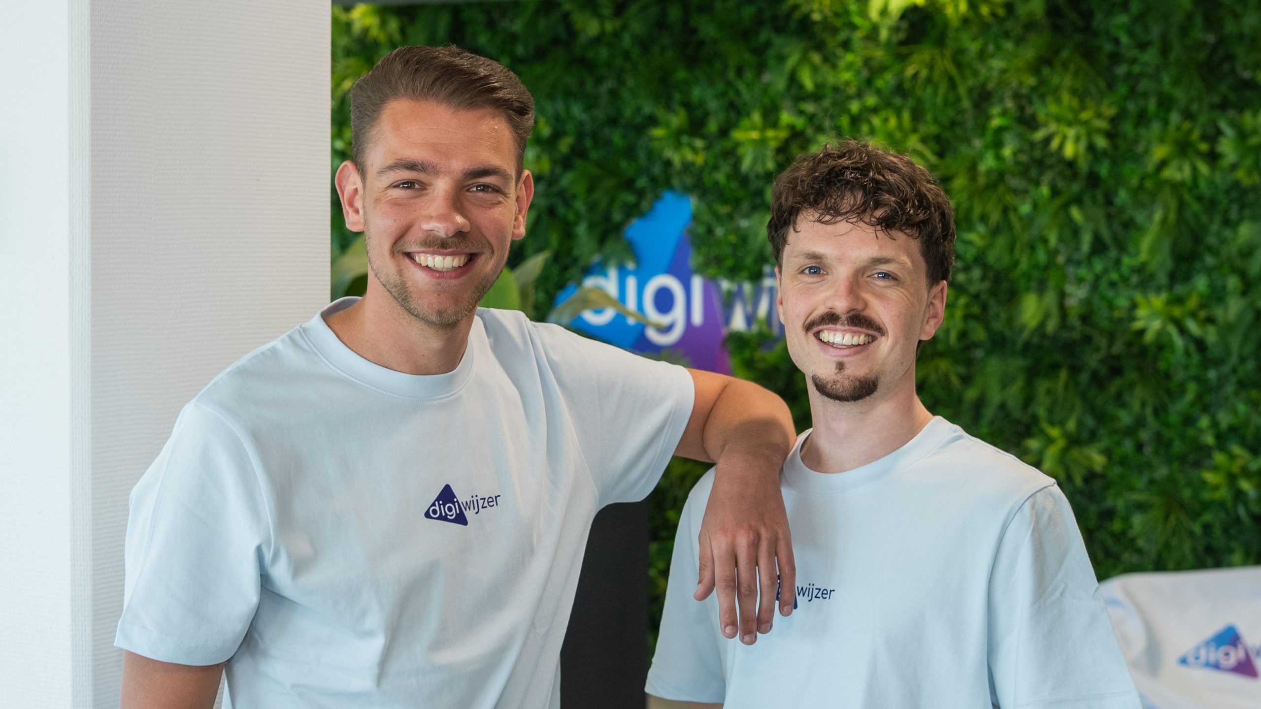 Twee Digiwijzer-collega's staan samen voor een groene plantenwand op kantoor en werken gezamenlijk aan digitale geletterdheid in het onderwijs.