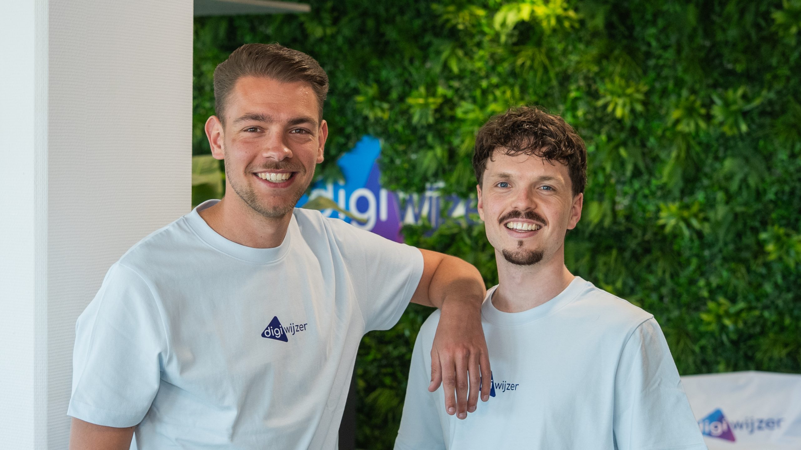 Twee Digiwijzer-medewerkers poseren samen voor de groene plantenwand op kantoor, als vertegenwoordigers van het team digitale geletterdheid.