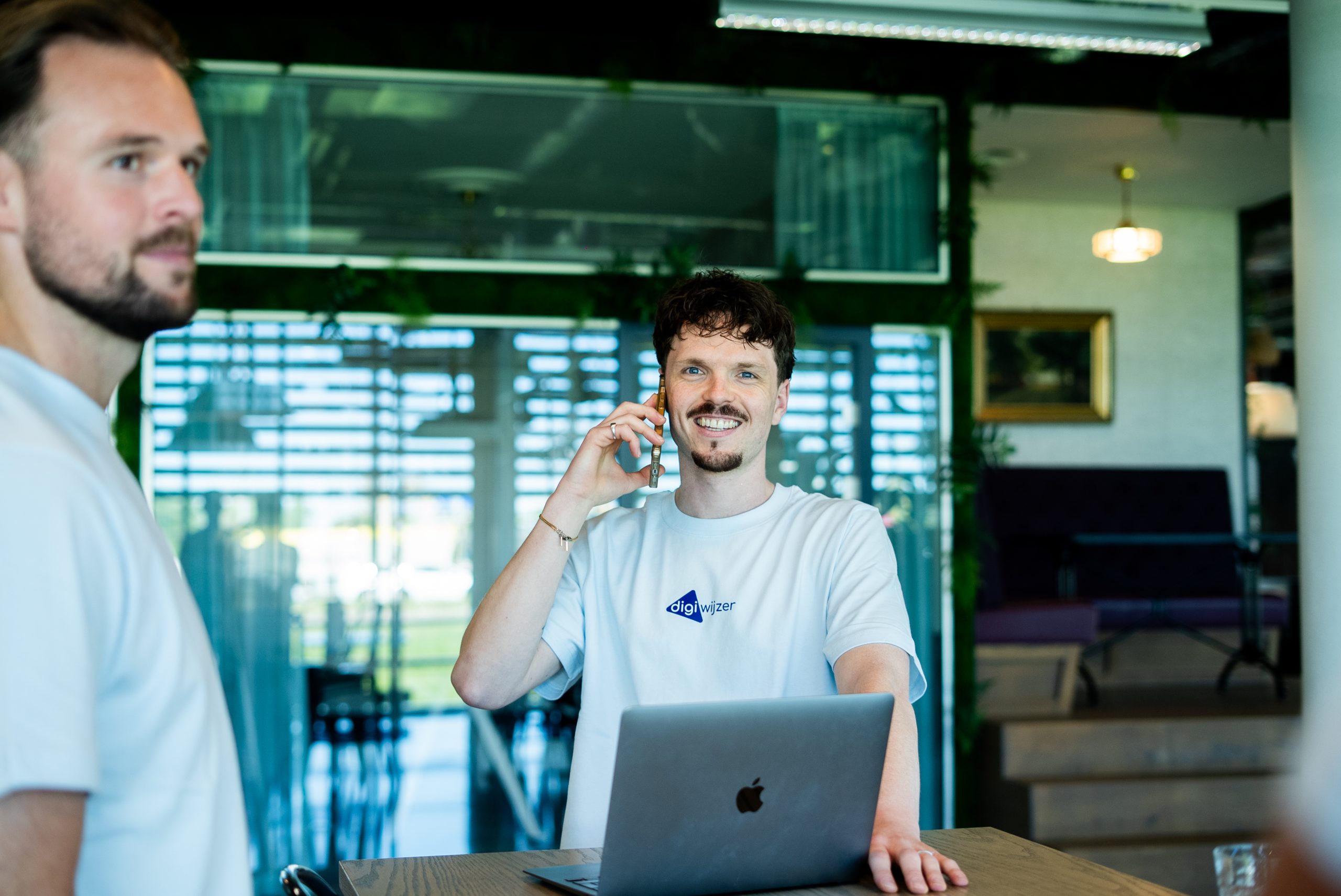 Een Digiwijzer-medewerker voert een telefoongesprek terwijl collega's samenwerken aan digitale geletterdheid op kantoor.
