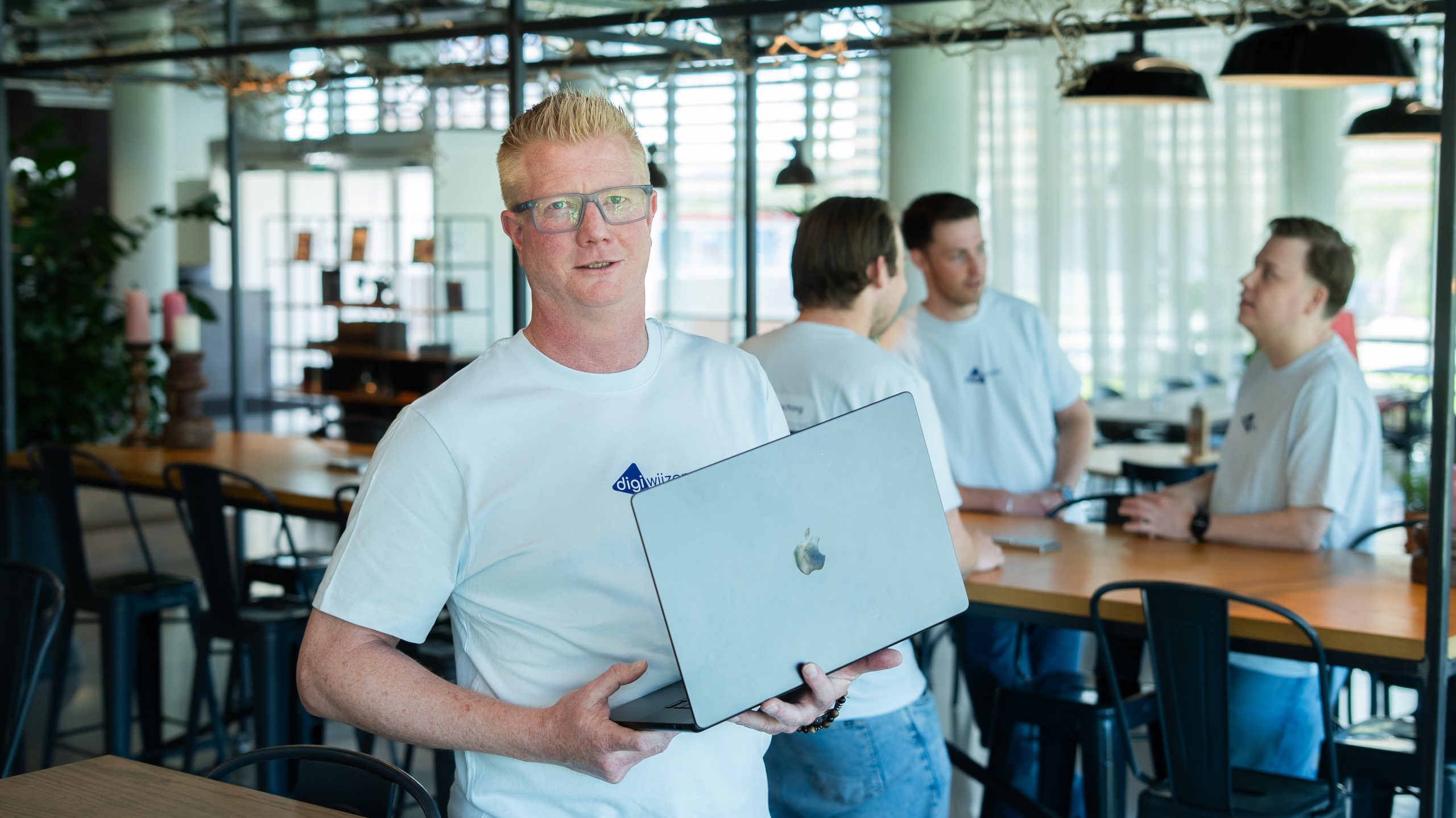 Digicoach met laptop terwijl collega's samenwerken op de achtergrond.