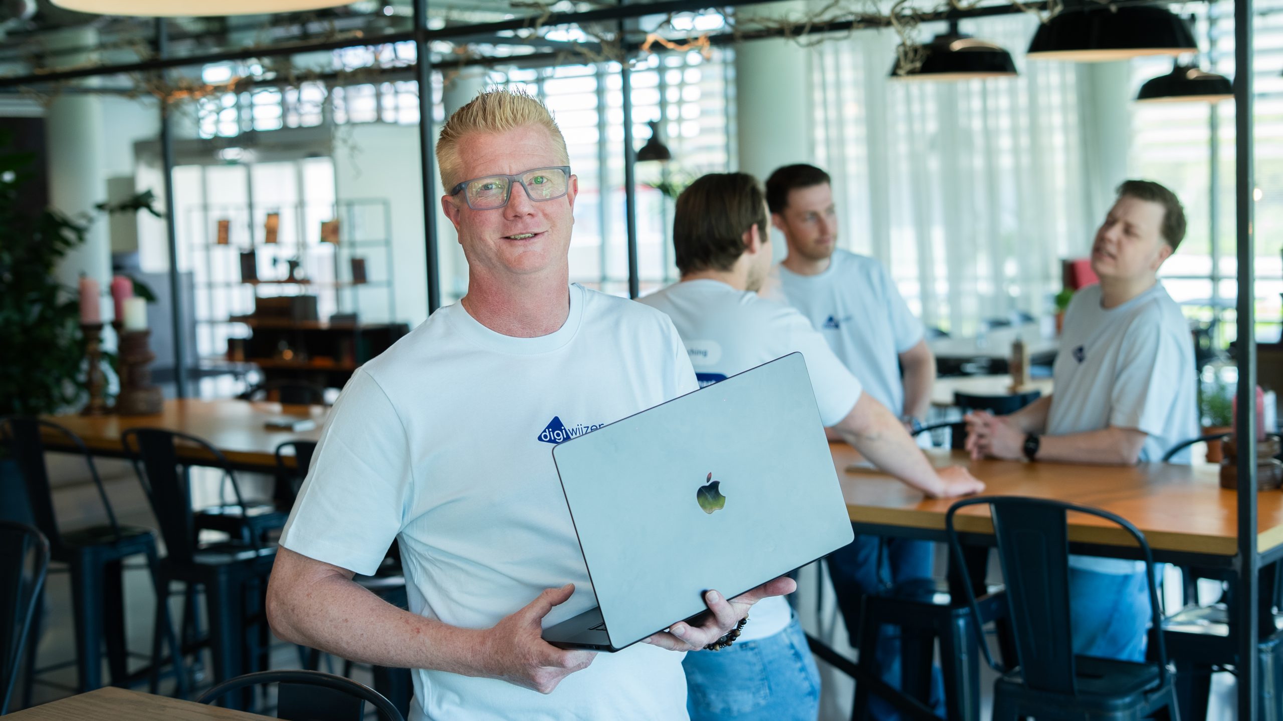 Medewerker van Digiwijzer houdt een laptop vast terwijl collega's op de achtergrond samenwerken.