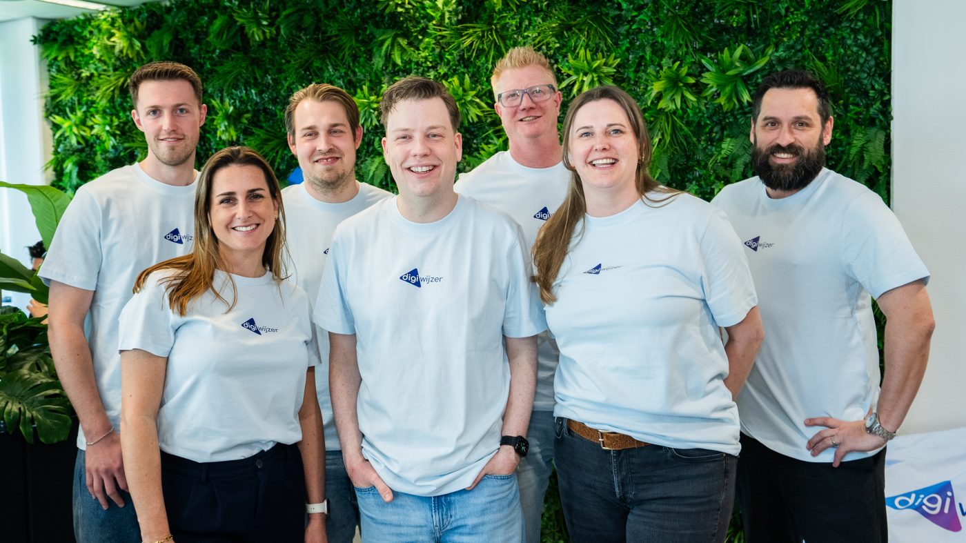 Team van Digiwijzer poseert gezamenlijk voor een groene plantenwand op kantoor.