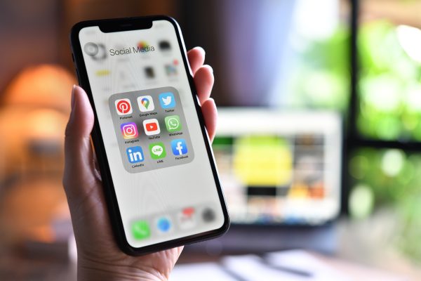 Hand houdt een smartphone vast met verschillende socialmedia-apps op het scherm, terwijl op de achtergrond een laptop zichtbaar is.