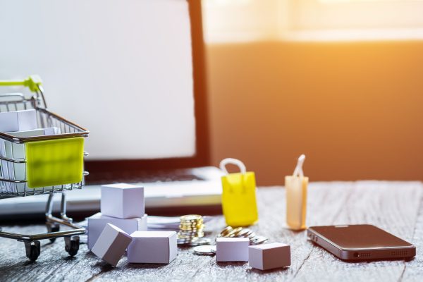 Winkelwagentje, smartphone, laptop en kleine pakketjes op een tafel als symbool voor online ondernemen en verkopen via digitale platforms.