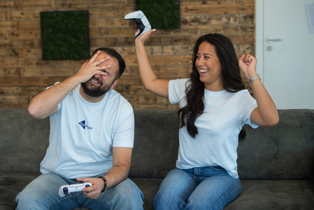 Twee volwassenen spelen samen een videogame op de bank. Eén persoon houdt lachend een gamecontroller omhoog, terwijl de ander met een glimlach zijn gezicht bedekt tijdens het gamen.