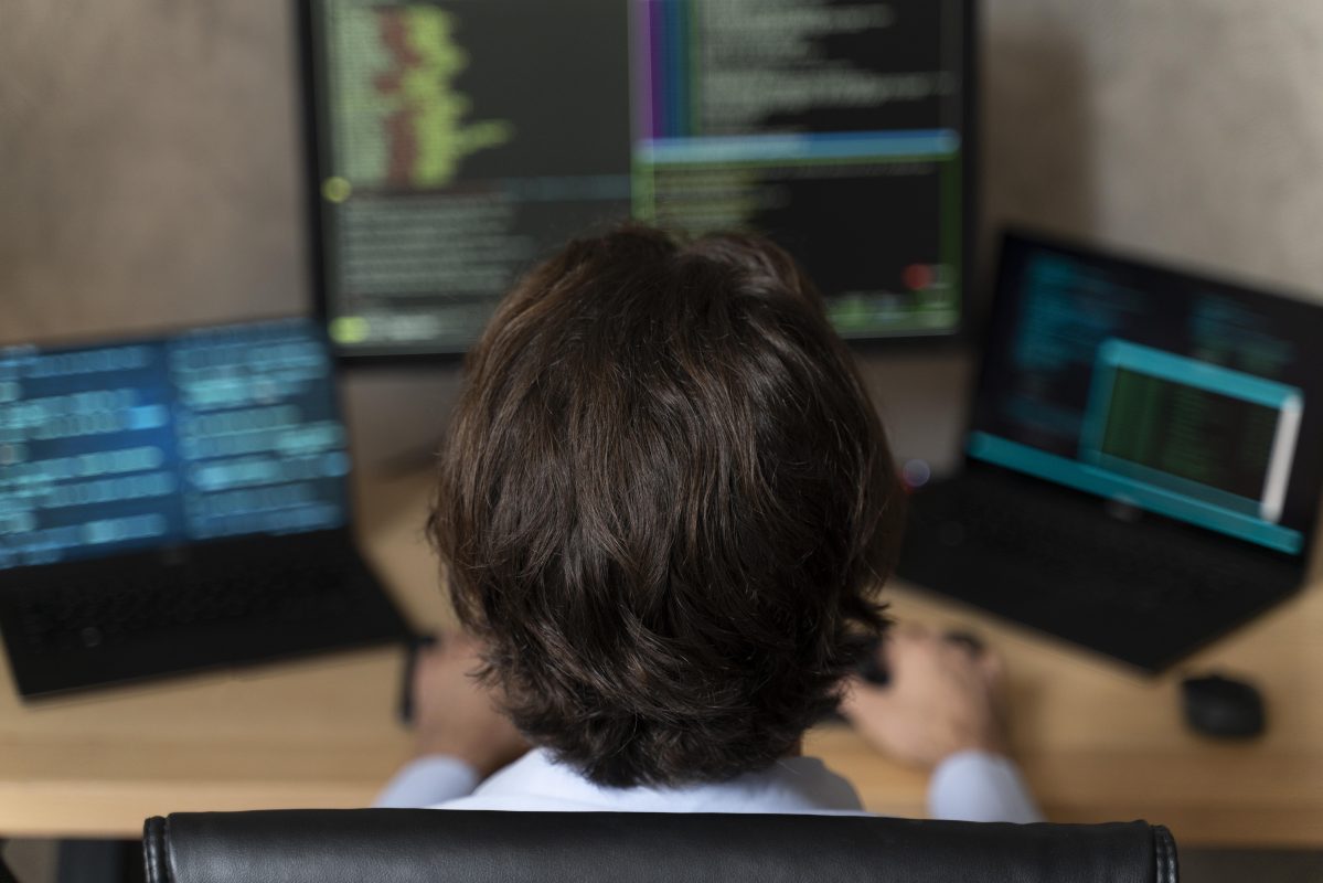 Jongere achter een computer met meerdere schermen waarop programmeercode te zien is.