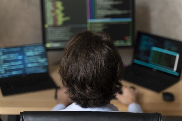 Jongere achter een computer met meerdere schermen waarop programmeercode te zien is.