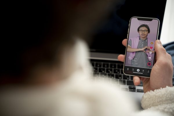 Persoon houdt een smartphone met de TikTok For You Page vast terwijl op de achtergrond een laptop zichtbaar is.