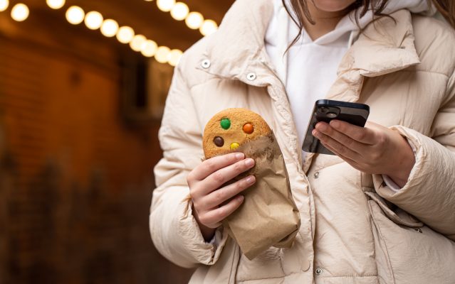 Jongere houdt een smartphone en een koekje vast bij een blog over dark patterns en online beïnvloeding.